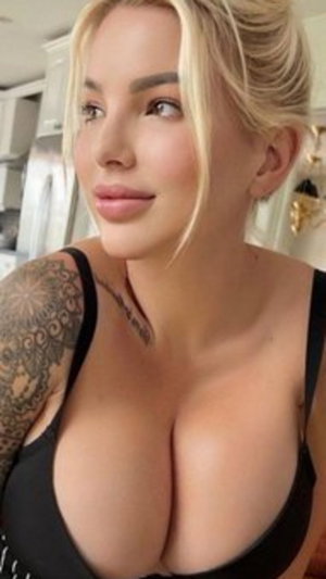 Blonde woman in black bra with tattooed arm, indoors in kitchen.