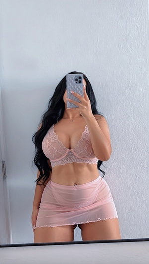 Woman in pink lace bra and sheer skirt taking mirror selfie indoors.