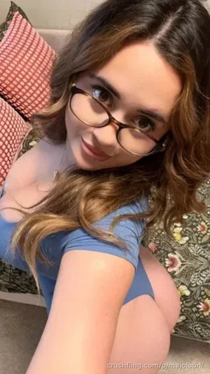 Woman with wavy brown hair and glasses in blue bodysuit, sitting on patterned couch indoors.