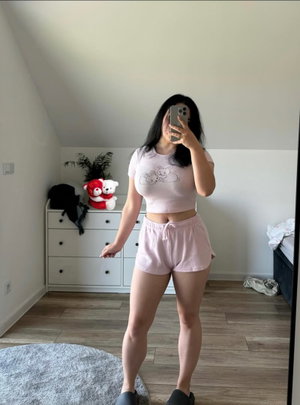 Woman in pink crop top and shorts takes mirror selfie in bedroom.
