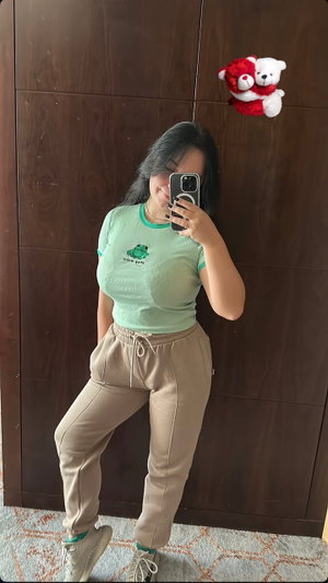 Woman in green t-shirt and beige pants taking mirror selfie in bedroom.