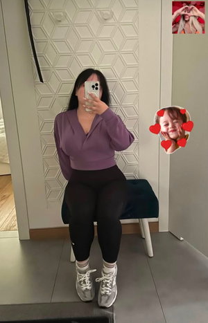 Woman in purple top and black leggings takes mirror selfie in bedroom.