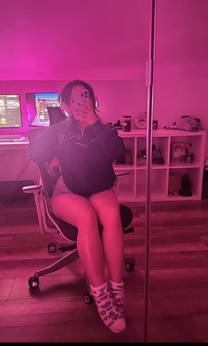 Woman in black hoodie and shorts sits in office chair, taking mirror selfie in pink-lit room.