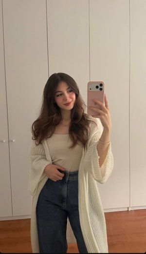 Woman with long brown hair in cream cardigan and jeans, taking mirror selfie indoors.