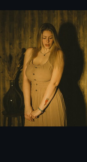 Woman in beige dress with buttons, standing indoors near wooden wall and vase.