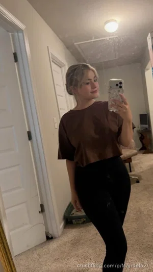 Woman in brown crop top and black pants taking mirror selfie in hallway.