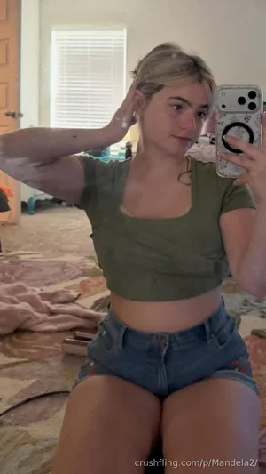 Woman in green crop top and denim shorts sits on bed, taking mirror selfie in bedroom.