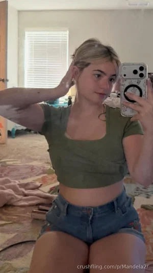 Woman in olive crop top and denim shorts taking mirror selfie in bedroom.