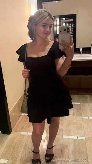 Woman in black dress with ruffled sleeves, holding phone, standing in tiled bathroom.