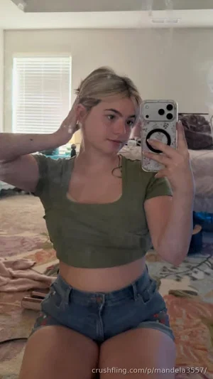 Woman in olive crop top and denim shorts sits on bed, holding phone for mirror selfie.