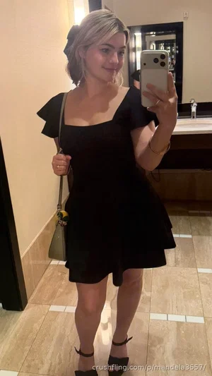 Woman in black dress with ruffled sleeves, holding phone, standing in tiled bathroom.