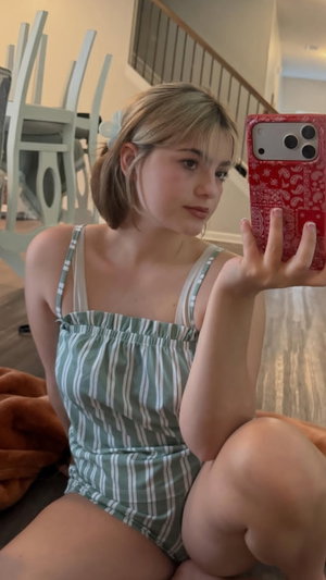 Woman in striped romper taking mirror selfie indoors, sitting on floor.