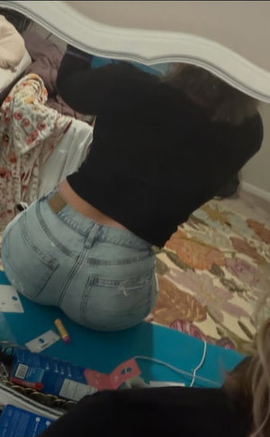 Woman in black top and jeans, kneeling on bed, mirror selfie, indoor setting.