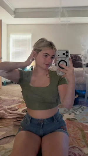 Woman in green crop top and denim shorts sits on bed, taking mirror selfie in bedroom.