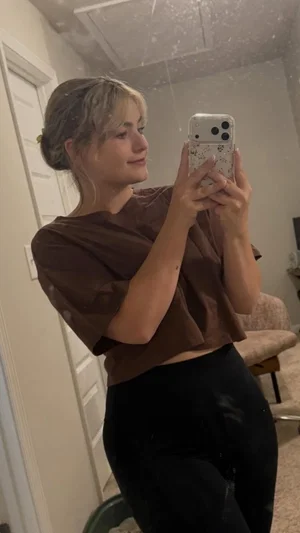 Woman with blonde hair in a brown crop top and black leggings taking a mirror selfie indoors.