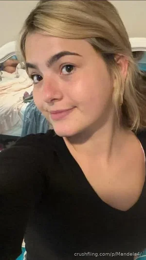 Woman with blonde hair in a black top, smiling in a bedroom selfie.