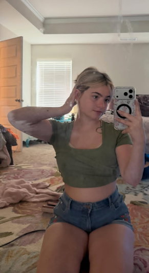 Blonde woman in green crop top and shorts taking bedroom selfie.