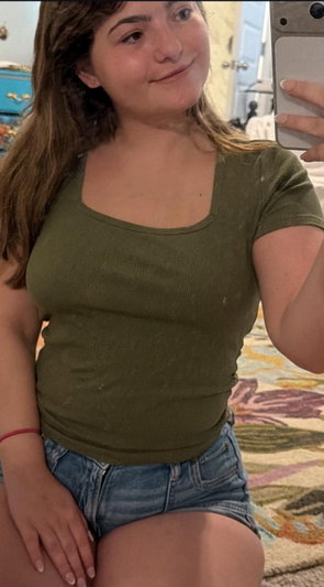 Brunette woman in green top and denim shorts taking selfie.