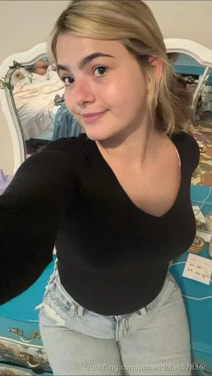 Woman with blonde hair in black top and jeans, posing in bedroom.