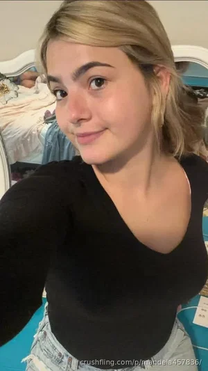 Woman with blonde hair in black top and jeans, taking selfie in bedroom.