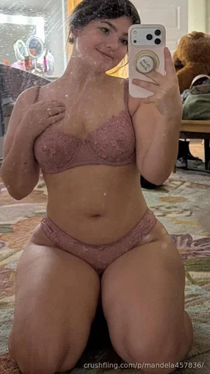 Woman in pink lace lingerie kneeling on carpet, holding phone for mirror selfie.