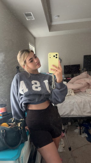 Woman in gray crop sweatshirt and black shorts takes mirror selfie in bedroom.