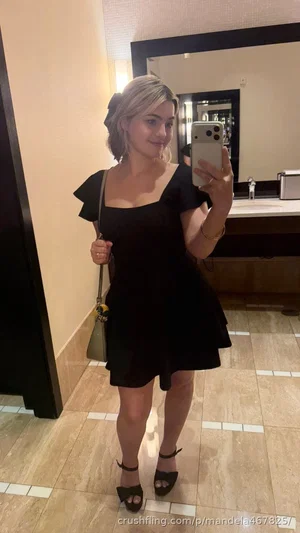 Woman in black dress taking mirror selfie in bathroom, holding phone and purse.