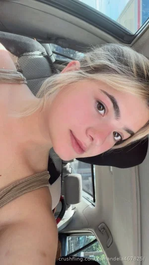 Woman with blonde hair in a car, wearing a strap top, looking at camera.