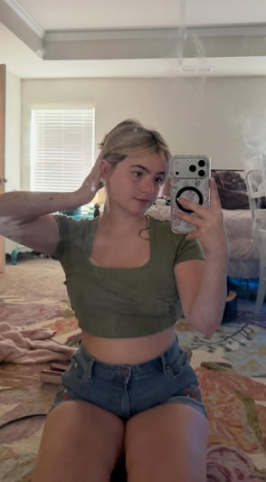 Blonde woman in green top and shorts taking mirror selfie in bedroom.