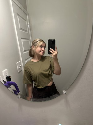 Woman with blonde hair in olive crop top and black pants, mirror selfie in indoor setting.
