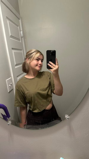 Woman with blonde hair in olive green crop top and black pants, taking mirror selfie indoors.