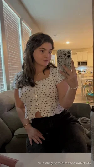 Woman in patterned crop top and black pants takes mirror selfie in living room.