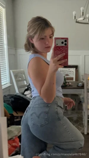 Woman in light blue tank top and jeans taking mirror selfie in home setting.
