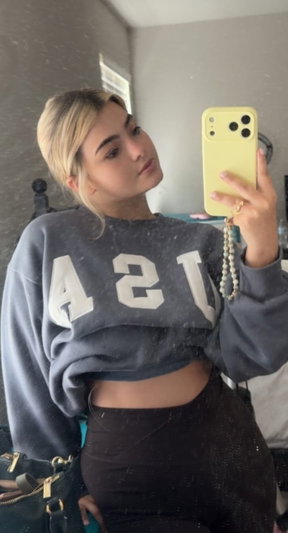 Woman with blonde hair in a gray crop sweatshirt and black pants taking a mirror selfie in a