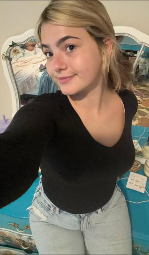 Woman with blonde hair in black top and jeans, taking selfie in bedroom.