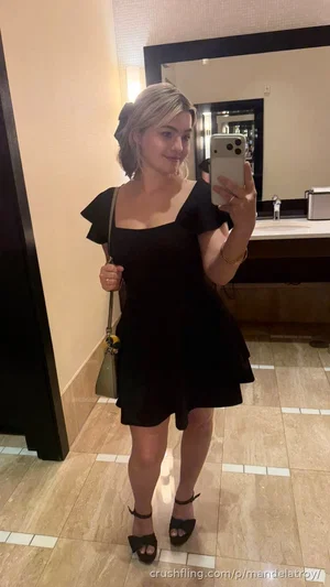 Woman in black dress taking mirror selfie in bathroom, holding phone and purse.