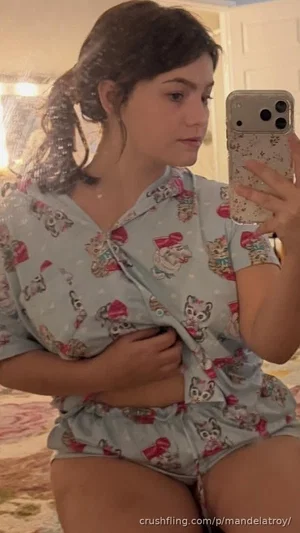 Woman in light blue pajama set with cartoon print, sitting on bed, holding phone for mirror selfie.