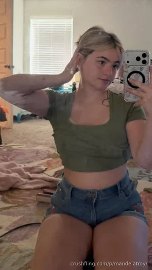 Woman in olive crop top and denim shorts sits on bed, holding phone for mirror selfie.