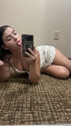 Woman in white polka-dot top lying on carpet, taking mirror selfie, tongue out.