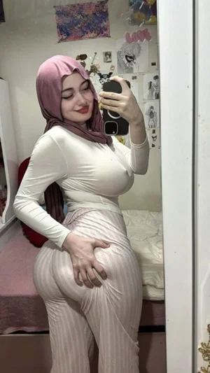 Woman in pink hijab and white outfit poses in mirror selfie in bedroom.