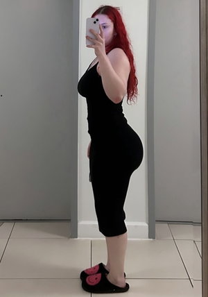 Red-haired woman in black dress taking mirror selfie indoors.