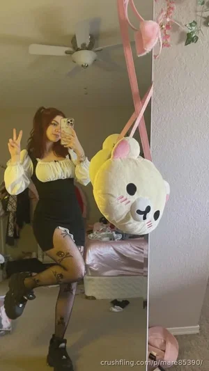 Woman in black dress with white ruffled sleeves, fishnet stockings, and tattoos poses in bedroom