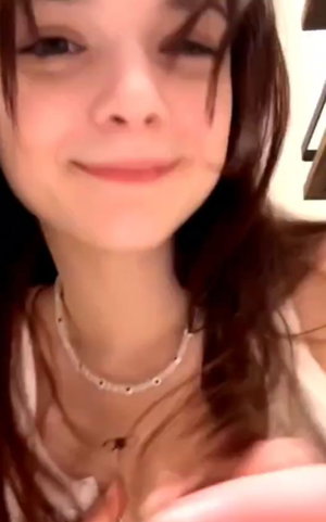 Woman with long brown hair wearing a light top and necklace, smiling in a close-up selfie.