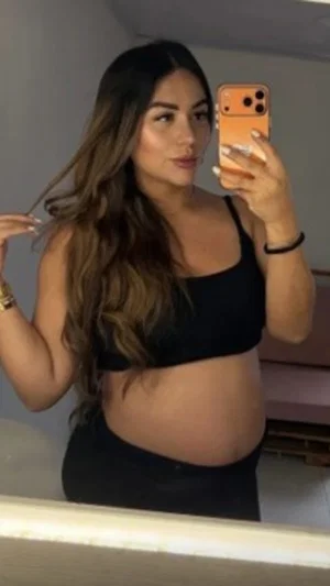 Woman with long brown hair in black crop top and pants, taking mirror selfie indoors.