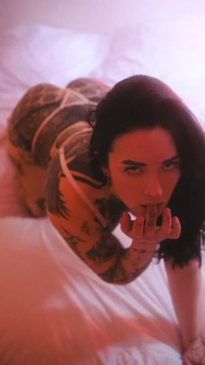 Woman with tattoos lying on bed, wearing white top, looking at camera.