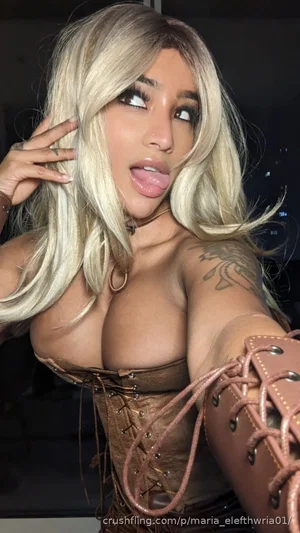 Woman with blonde hair, tattoos, wearing brown corset and arm cuffs, posing indoors.