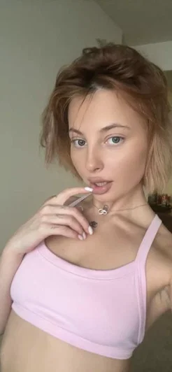 Woman with short brown hair in pink crop top, holding cigarette indoors.