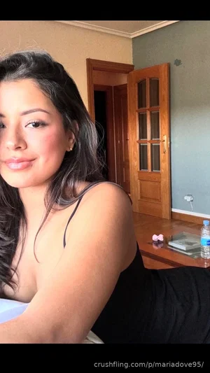 Woman with dark hair in black top posing indoors, partial view of room with wooden door.