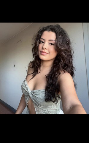 Woman with curly dark hair in a light-colored top, taking a selfie indoors.