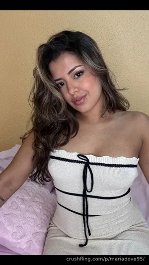 Woman with wavy hair in white strapless top sitting on pink bedding indoors.